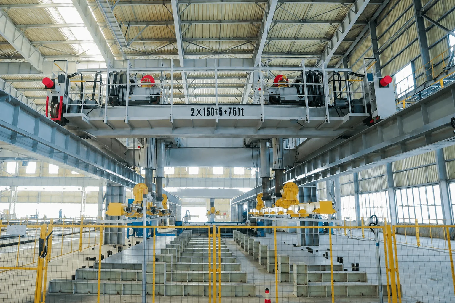 Grey overhead crane with paired handling attachments above a guarded steel-processing line