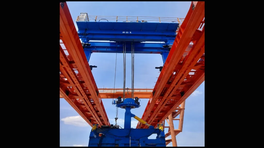 Crane installation showing multiple wire rope falls and a hook block below a blue lifting assembly between orange girders