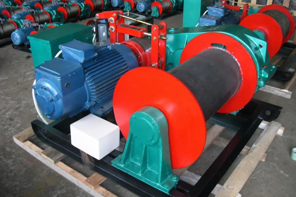 Base-frame electric winch in a factory with blue motor, red drum flanges, gearbox and visible brake assembly