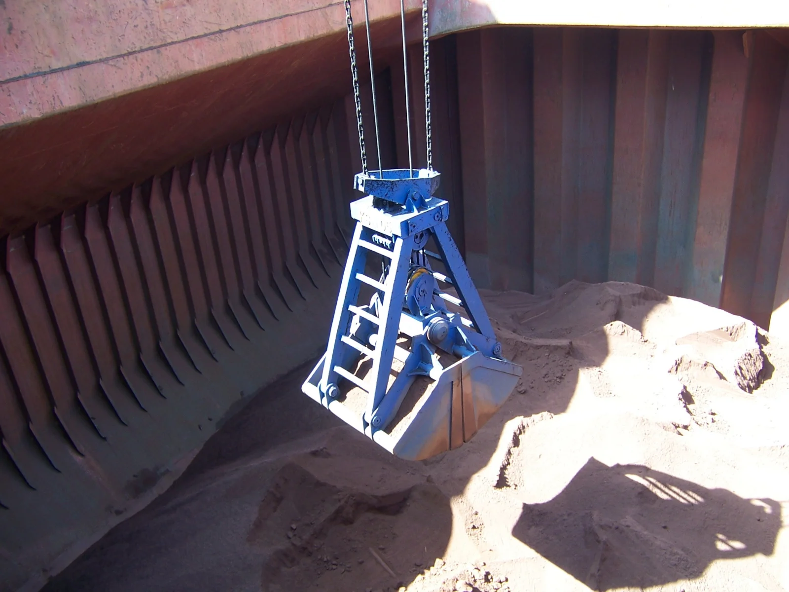 Rope-operated blue clamshell grab handling bulk material inside a ship hold