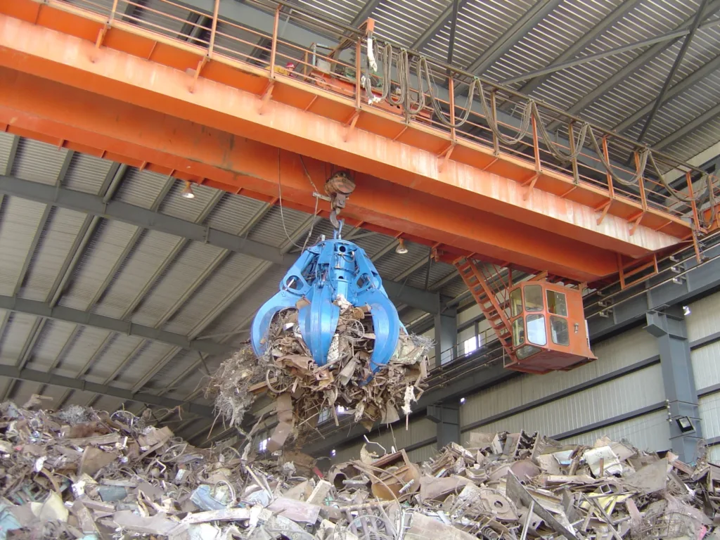 Blue multi-tine grab carrying scrap metal under a bridge crane