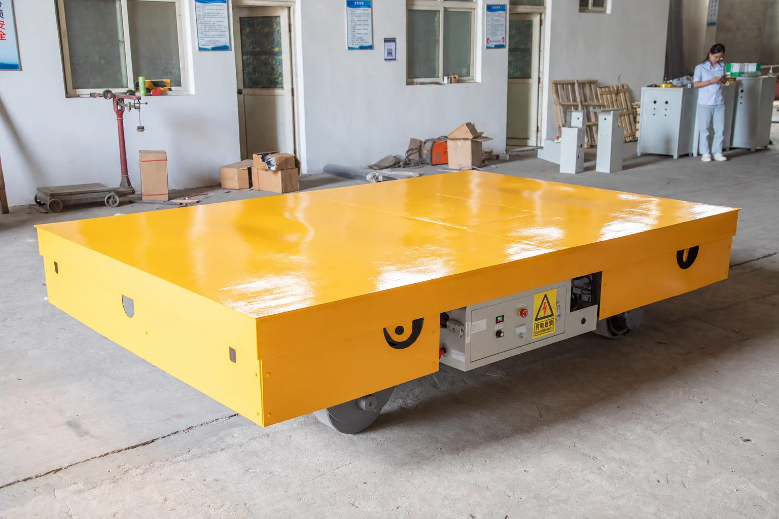 Yellow battery-powered trackless flat-deck cart with visible floor-contact wheels and side control enclosure