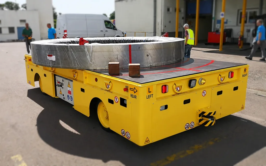 Yellow wheeled transfer cart carrying a large steel ring outside a workshop
