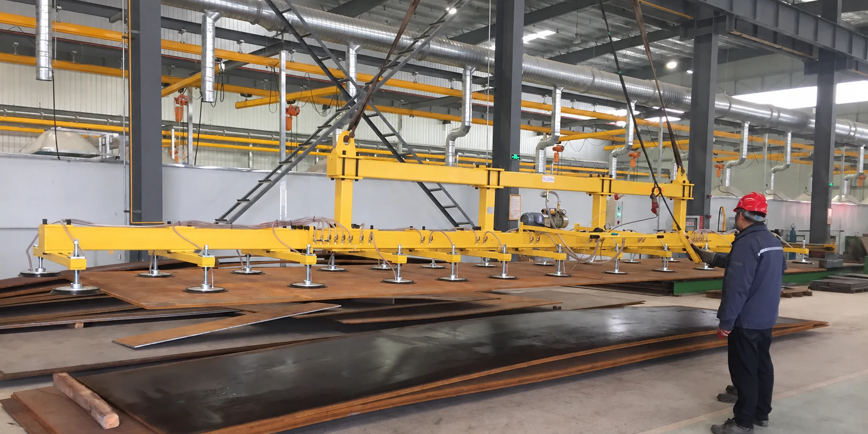 Long vacuum lifting beam with distributed suction pads over steel plates in a factory